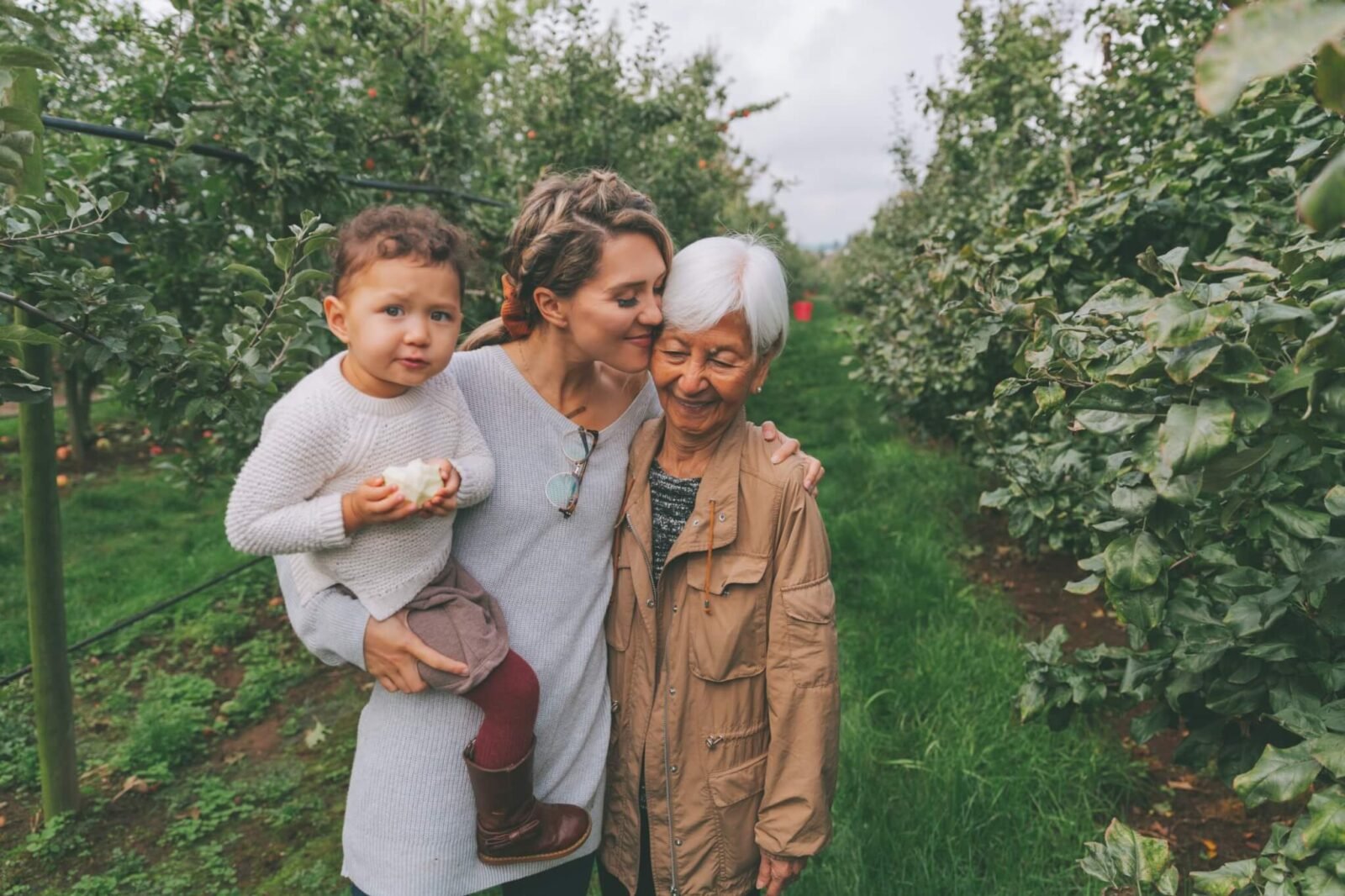 A woman is seen in an orchard, holding her son. She has her arm around her mother.