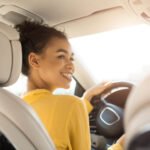 A Black woman is pictured driving a car. It appears as though the person in the back seat is taking the photo. She is wearing a yellow sweater and smiling.