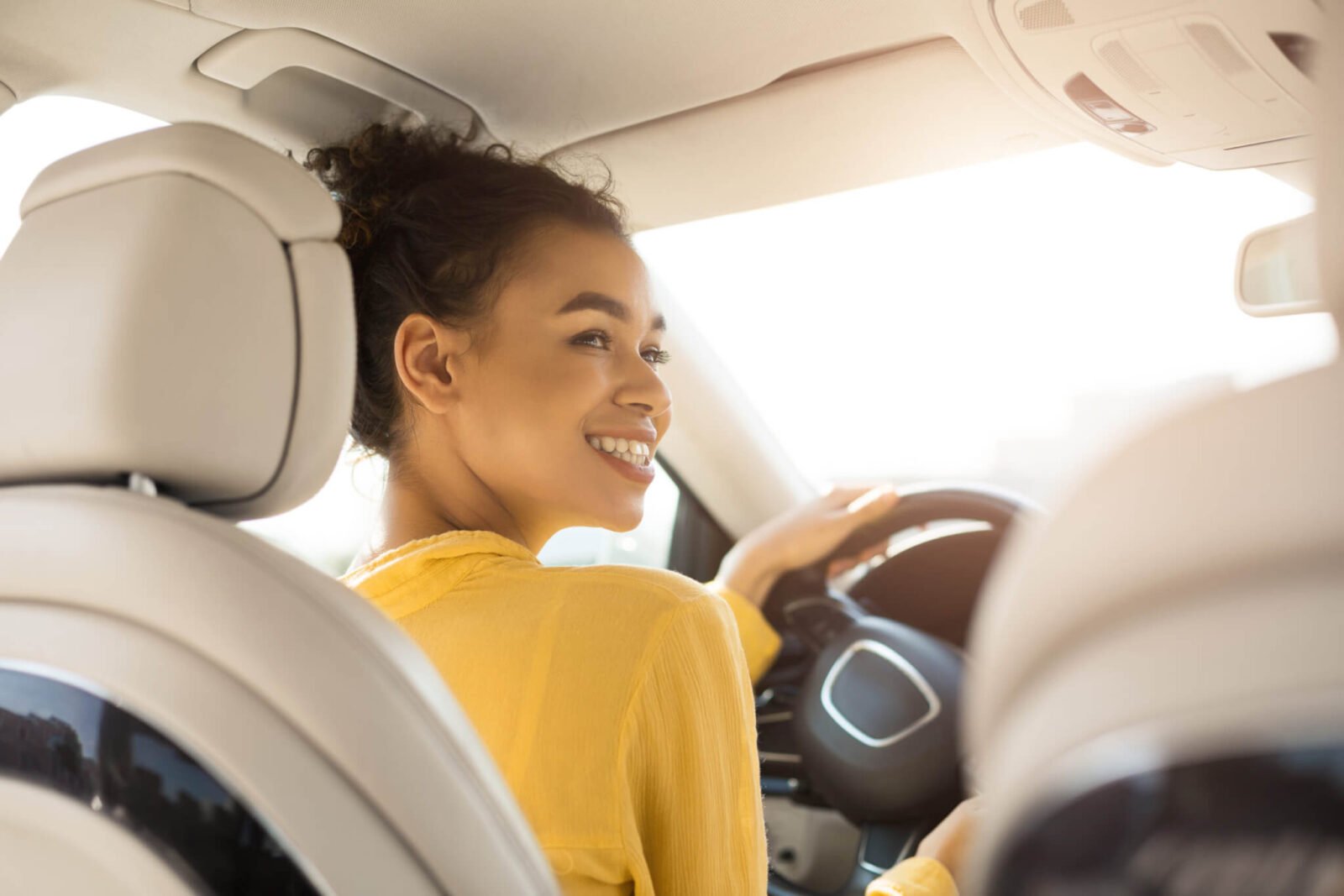 A Black woman is pictured driving a car. It appears as though the person in the back seat is taking the photo. She is wearing a yellow sweater and smiling.
