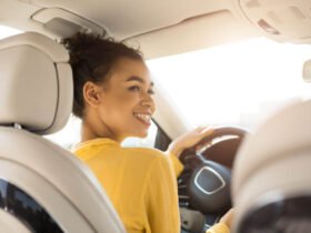 A Black woman is pictured driving a car. It appears as though the person in the back seat is taking the photo. She is wearing a yellow sweater and smiling.