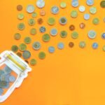 A glass jar is laying on a table with coins spilling out of it. The table is orange.