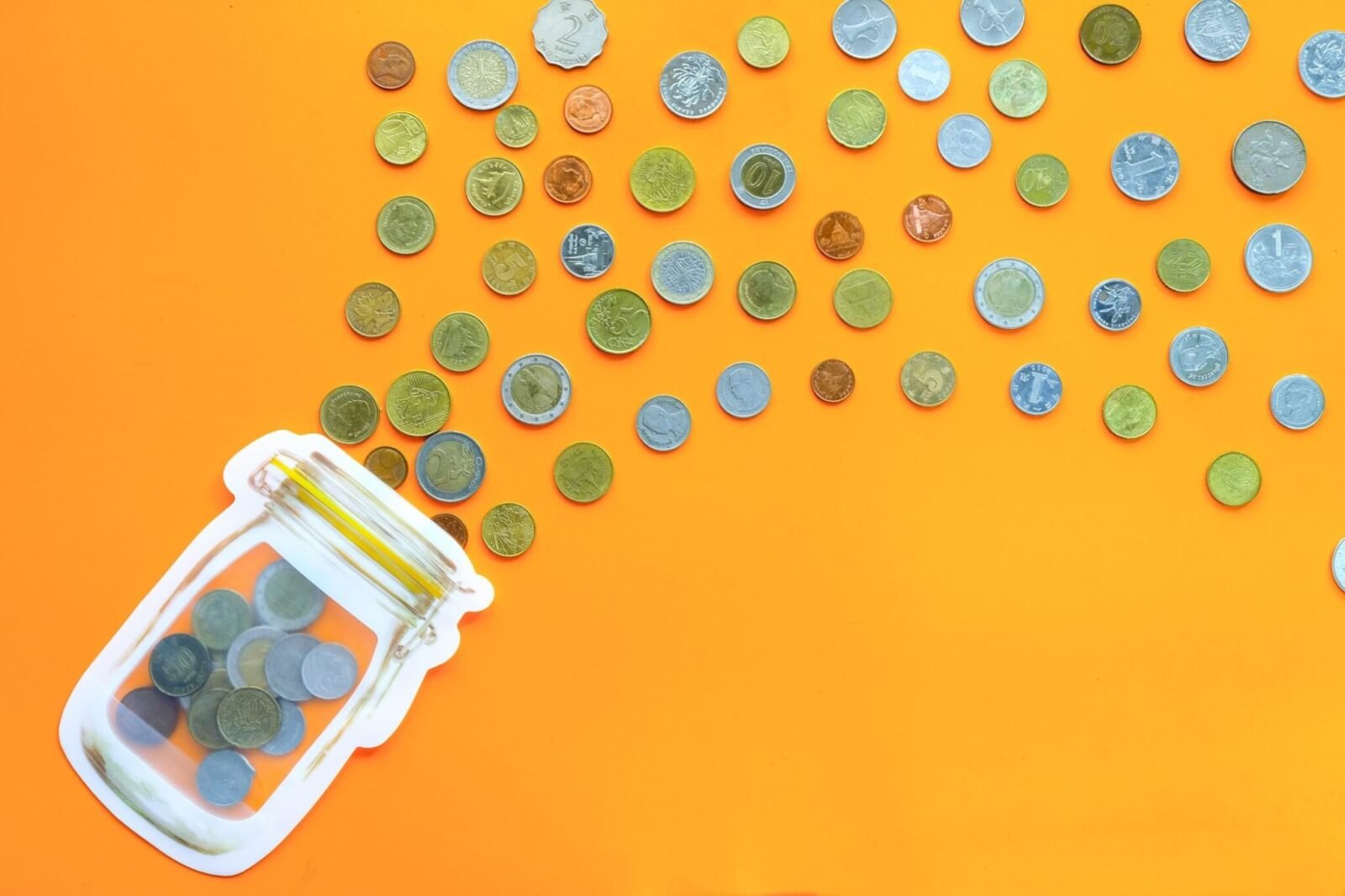 A glass jar is laying on a table with coins spilling out of it. The table is orange.