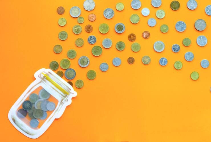 A glass jar is laying on a table with coins spilling out of it. The table is orange.
