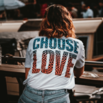 Woman sitting at a bar with her back to the camera. It's modern day, but she's wearing 80s style jeans with a white shirt tucked in. On the back of her shirt is blue and orange checkered text reading 'Choose Love'.