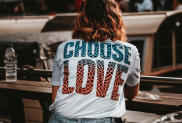 Woman sitting at a bar with her back to the camera. It's modern day, but she's wearing 80s style jeans with a white shirt tucked in. On the back of her shirt is blue and orange checkered text reading 'Choose Love'.