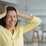 A woman is in a yellow top with her hands on her head. She appears stressed.