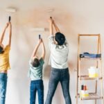 An adult and two children are seen painting a wall.