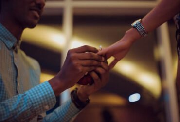 Man putting a ring on a woman's finger as he proposes on one knee. set against a backdrop of windows at night with lights reflected in the window.