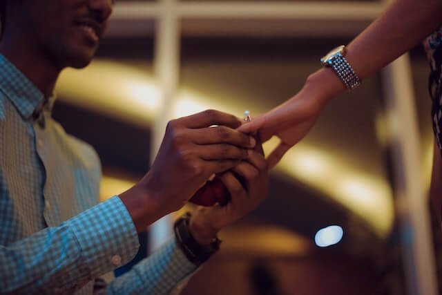 Man putting a ring on a woman's finger as he proposes on one knee. set against a backdrop of windows at night with lights reflected in the window.