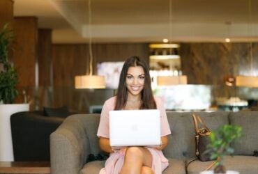 Woman with brown hair in a pink dress smiling at the camera looking over her laptop. She is sitting in a modern office.