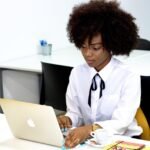 A woman wearing a white collared shirt works on an Apple laptop in an office. She chooses a new health insurance plan.