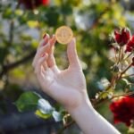 Image of woman's hand with red nail polish holding a physical representation of Bitcoin against the backdrop of a rose garden in full bloom.