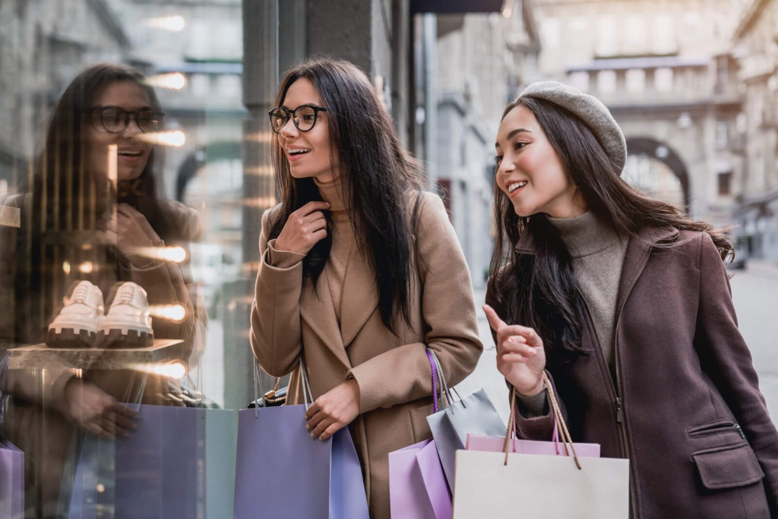 Two women are seen on a city street, looking in a store window at a pair of shoes. They are carrying shopping bags.