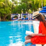 A woman in a red bathing suit cover-up is sitting on the side of a hotel pool. She is reading a book.