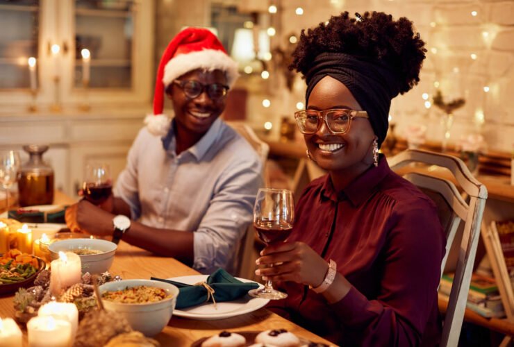 A woman sits at a table during a holiday party. The table is lit with candles and twinkle lights. She holds a glass of wine alongside her friend, who is wearing a Santa hat. They are filled with holiday cheer.