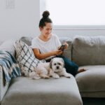 A woman wearing jeans and a white top and her small fluffy dog sit on a gray sectional. She is looking at her phone screen and smiling.