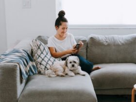 A woman wearing jeans and a white top and her small fluffy dog sit on a gray sectional. She is looking at her phone screen and smiling.