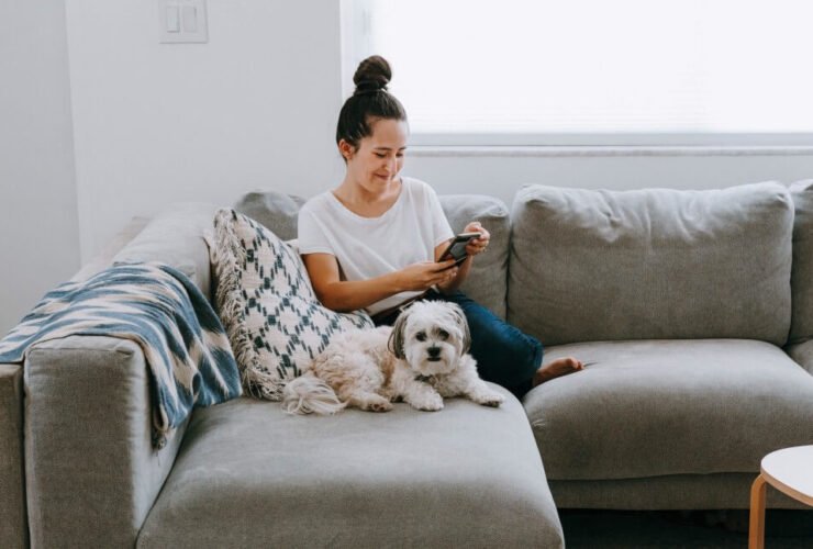 A woman wearing jeans and a white top and her small fluffy dog sit on a gray sectional. She is looking at her phone screen and smiling.