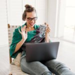 A woman sits at a laptop. She looks excited. She has brown hair and is wearing glasses.