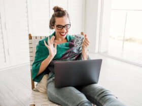 A woman sits at a laptop. She looks excited. She has brown hair and is wearing glasses.