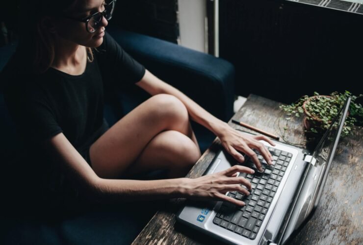 A woman types out business email examples on her laptop. She is wearing a dark t-shirt and glasses.