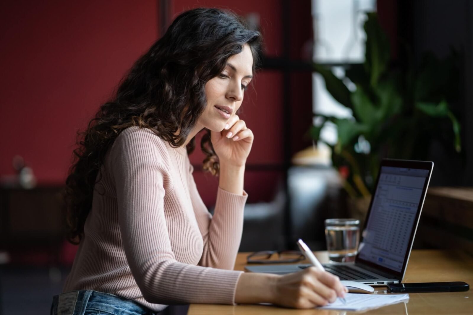 A woman with long hair in a pink shirt sits at a desk in front of a laptop. She is writing on a notepad.