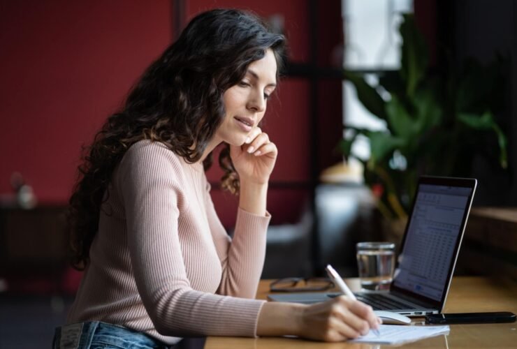A woman with long hair in a pink shirt sits at a desk in front of a laptop. She is writing on a notepad.