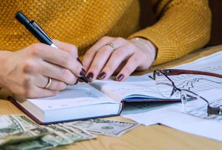 This is a close-up of a woman writing in a notebook. Her nails are painted and she is wearing rings. There is cash near the notebook, as well as a pair of glasses.