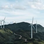 Nine wind turbines set along a windy road through a green mountain pass.