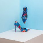 Pair of blue Aldo high heeled shoes with floral print. One is resting on a large white block. The other is hovering above against a light blue wall.