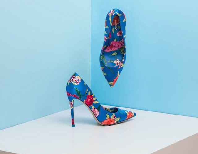 Pair of blue Aldo high heeled shoes with floral print. One is resting on a large white block. The other is hovering above against a light blue wall.