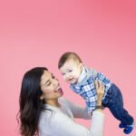 woman holding up her infant/toddler son against a pink background