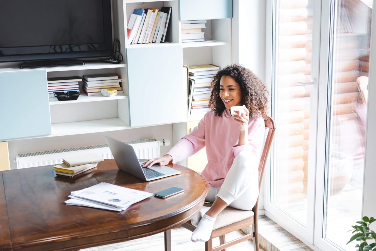 A Black woman is sitting at a table, sipping coffee and working on her laptop. It appears to be a sunny day, with light coming in behind her via a sliding glass door.