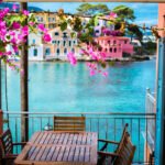 A table on a balcony is pictured. The balcony looks out on to a very blue body of water. There are also pink flowers on top of the balcony.
