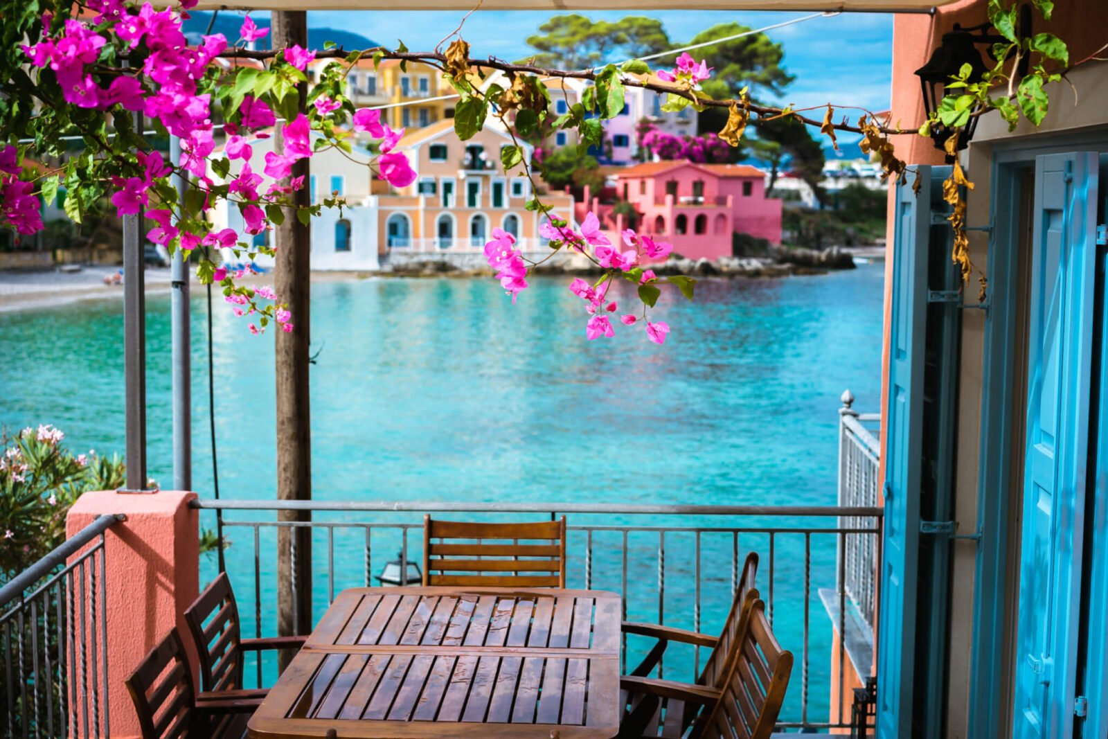 A table on a balcony is pictured. The balcony looks out on to a very blue body of water. There are also pink flowers on top of the balcony.