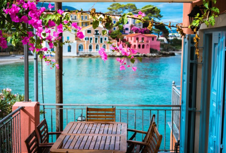 A table on a balcony is pictured. The balcony looks out on to a very blue body of water. There are also pink flowers on top of the balcony.