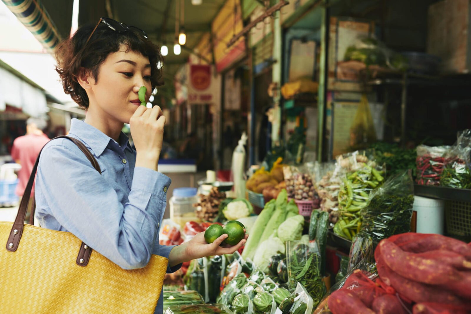 A woman wearing a blue shirt shops at an outdoor farmer's market, carrying a yellow tote bag.