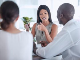 A black woman is pictured talking to two individuals. They are sitting at a table. She appears to be explaining something to them.