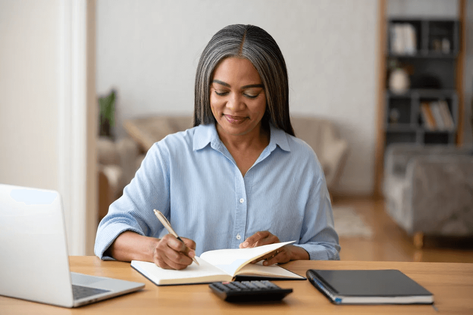 A woman sits at a desk and writes a budget in a notebook. She is Budgeting for Telehealth expenses. She has on a blue shirt and uses a computer as well.