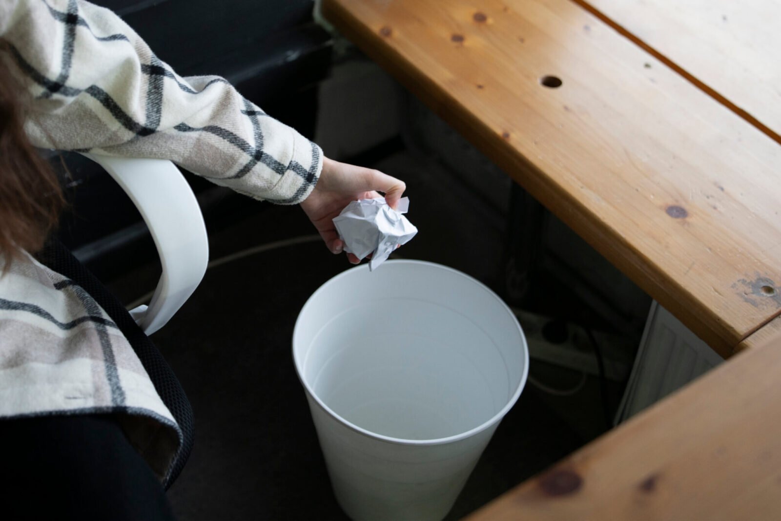 This is an image of a woman's hand throwing out a crumpled up piece of paper into a white trash can.