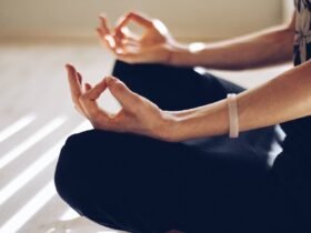 A woman sits cross legged, palms up and index finer and thumbs connecting. She appears to be meditating.