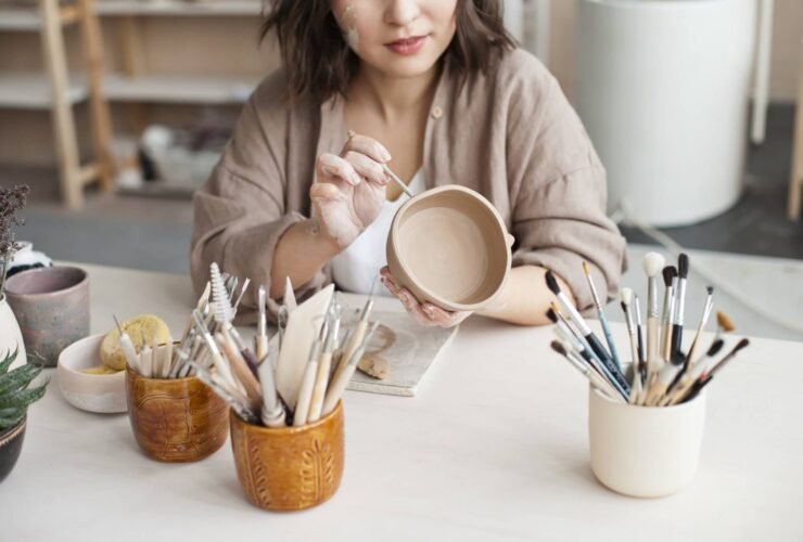 A woman sits at a table with a cup in her hand. There are other cups full of paintbrushes, and she is using one of them to decorate her cup.