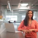 woman in a peach shirt with long grey hair standing in a white open space office next to a raised table with a laptop