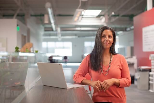 woman in a peach shirt with long grey hair standing in a white open space office next to a raised table with a laptop