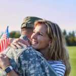 A woman with blonde hair is pictured hugging a man in a military uniform. She is holding an American flag.