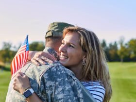 A woman with blonde hair is pictured hugging a man in a military uniform. She is holding an American flag.