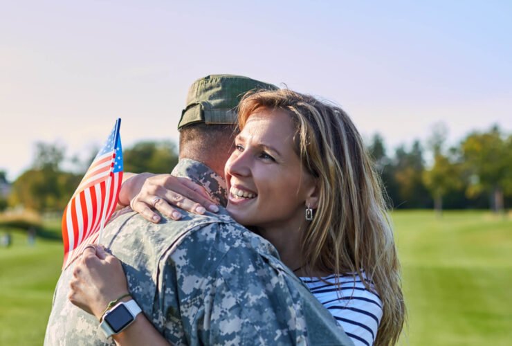 A woman with blonde hair is pictured hugging a man in a military uniform. She is holding an American flag.