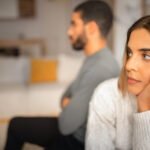 A woman is seen with her head in her hands, looking off in the distance. A man is behind her, with his arms crossed.