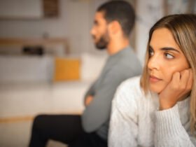 A woman is seen with her head in her hands, looking off in the distance. A man is behind her, with his arms crossed.