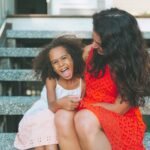 A Black woman is sitting on steps with a Black child. The woman is wearing a red dress. The child is wearing a white and pink dress. They are embracing and laughing.
